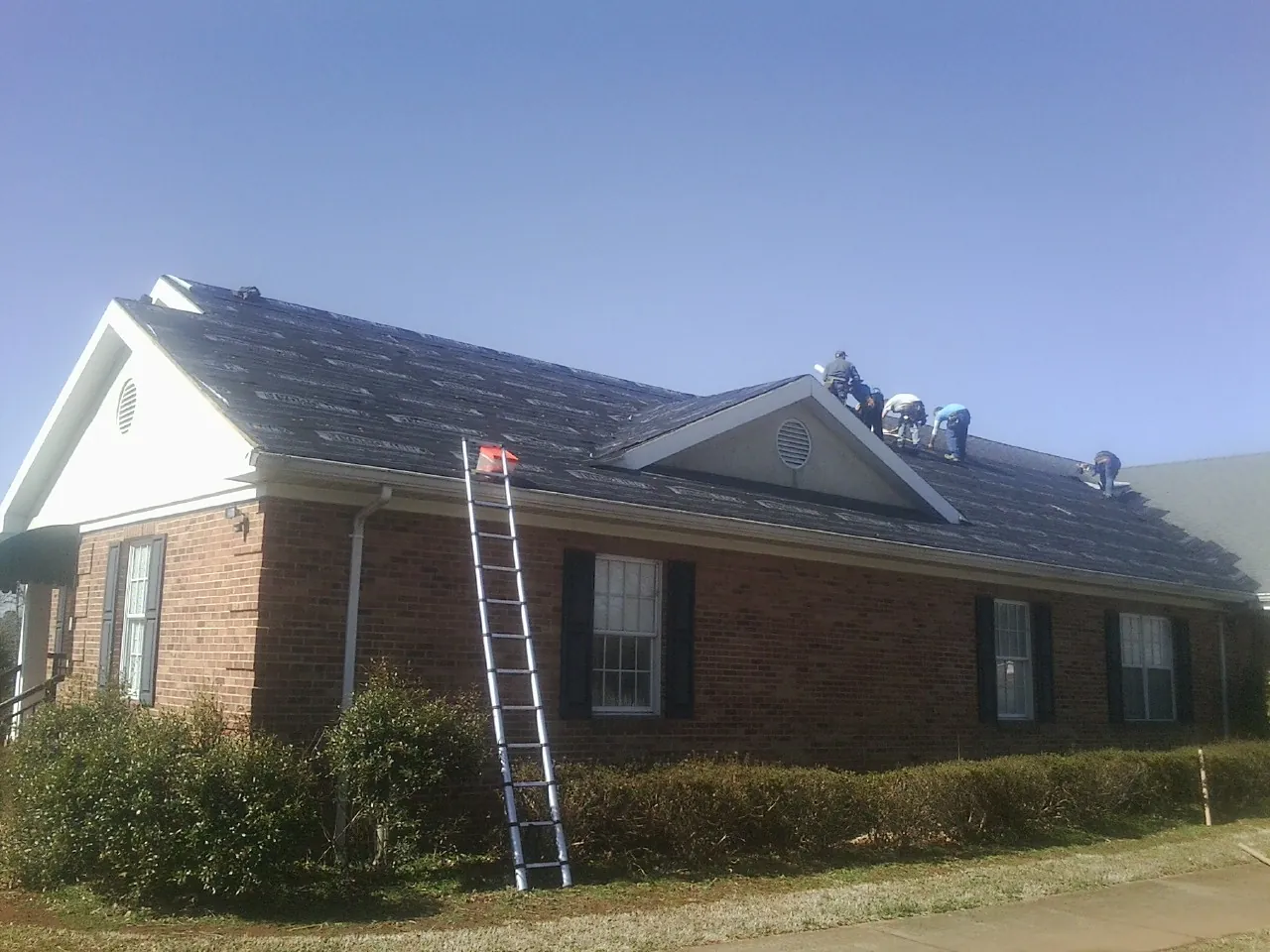 Crew tearing shingles off a brick ranch roof with a ladder set — Emergency Roof Repair work by our crew in Friesland