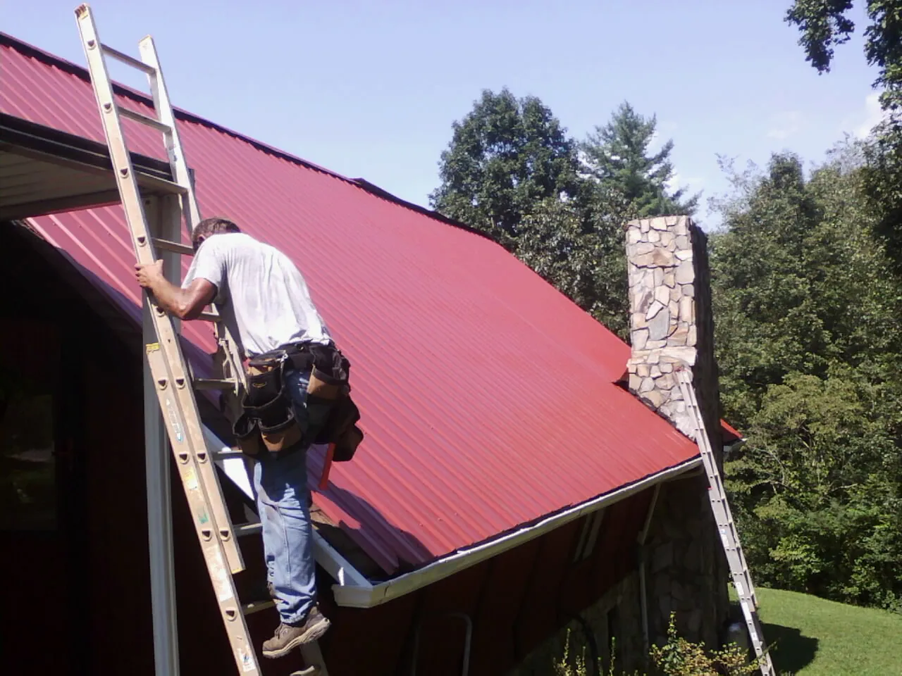 Roofer on a ladder beside a red metal roof and stone chimney — Soffit & Fascia Repair by Roofing Brothers in Friesland